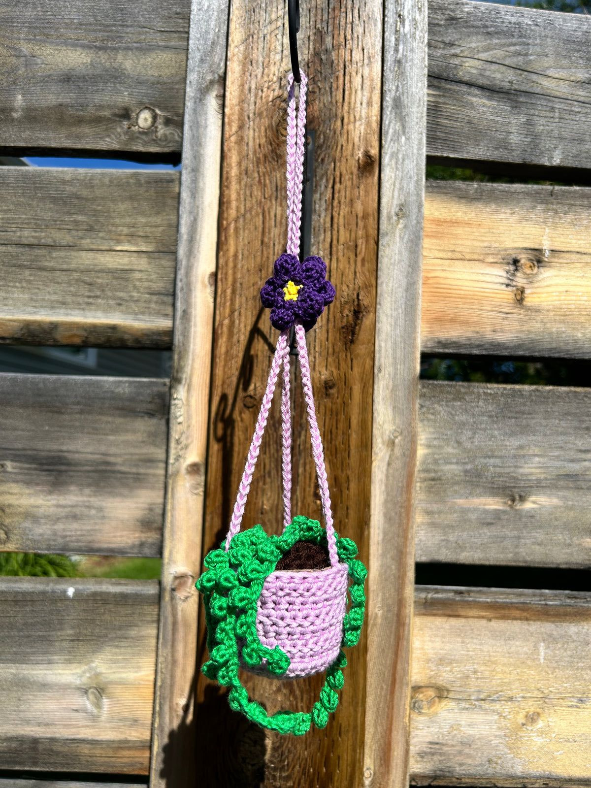 Crochet hanging plant holder with black cord and purple flower, photographed outdoors in a garden
